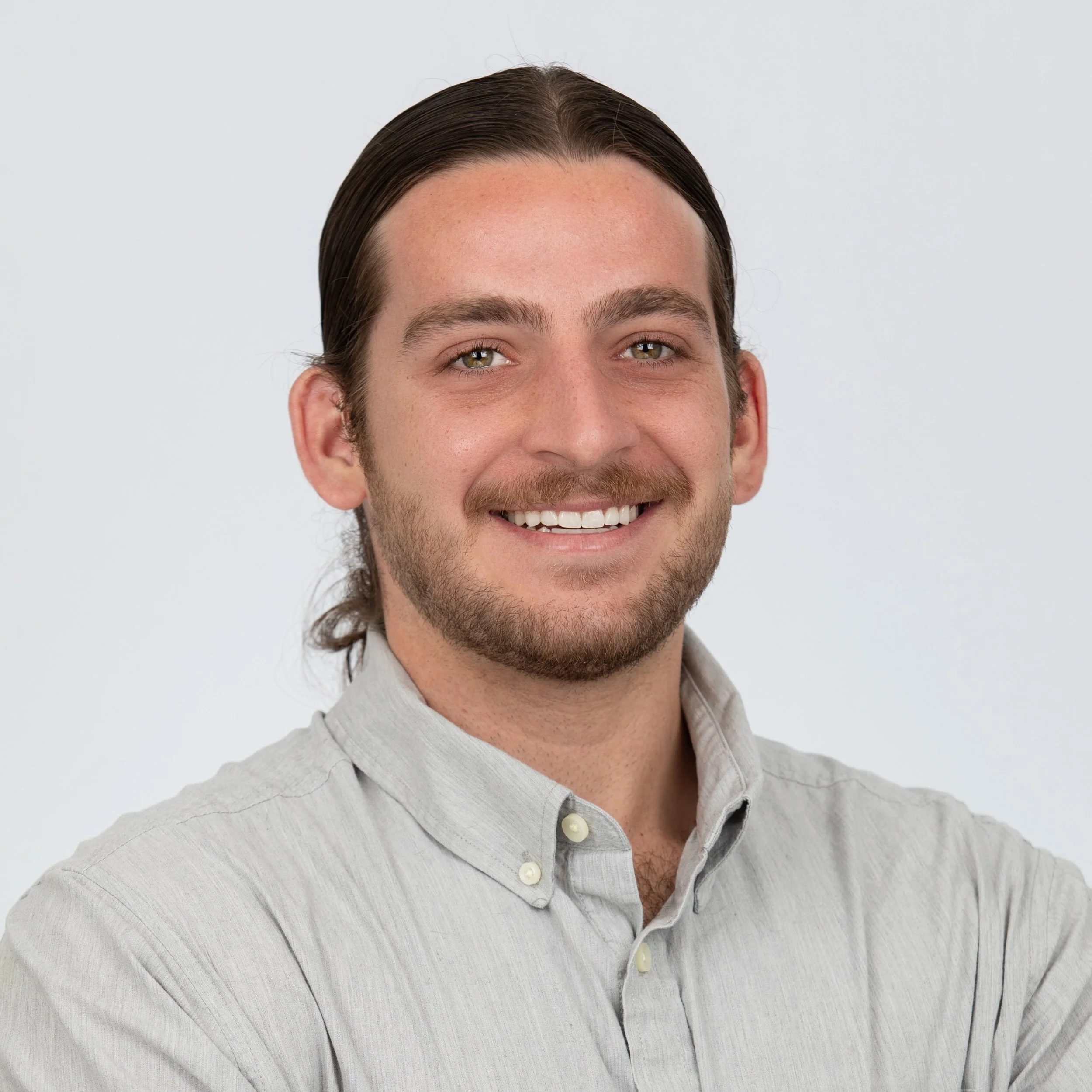 Smiling man with long brown hair pulled back in low ponytail and beard, wearing grey button up collared shirt.