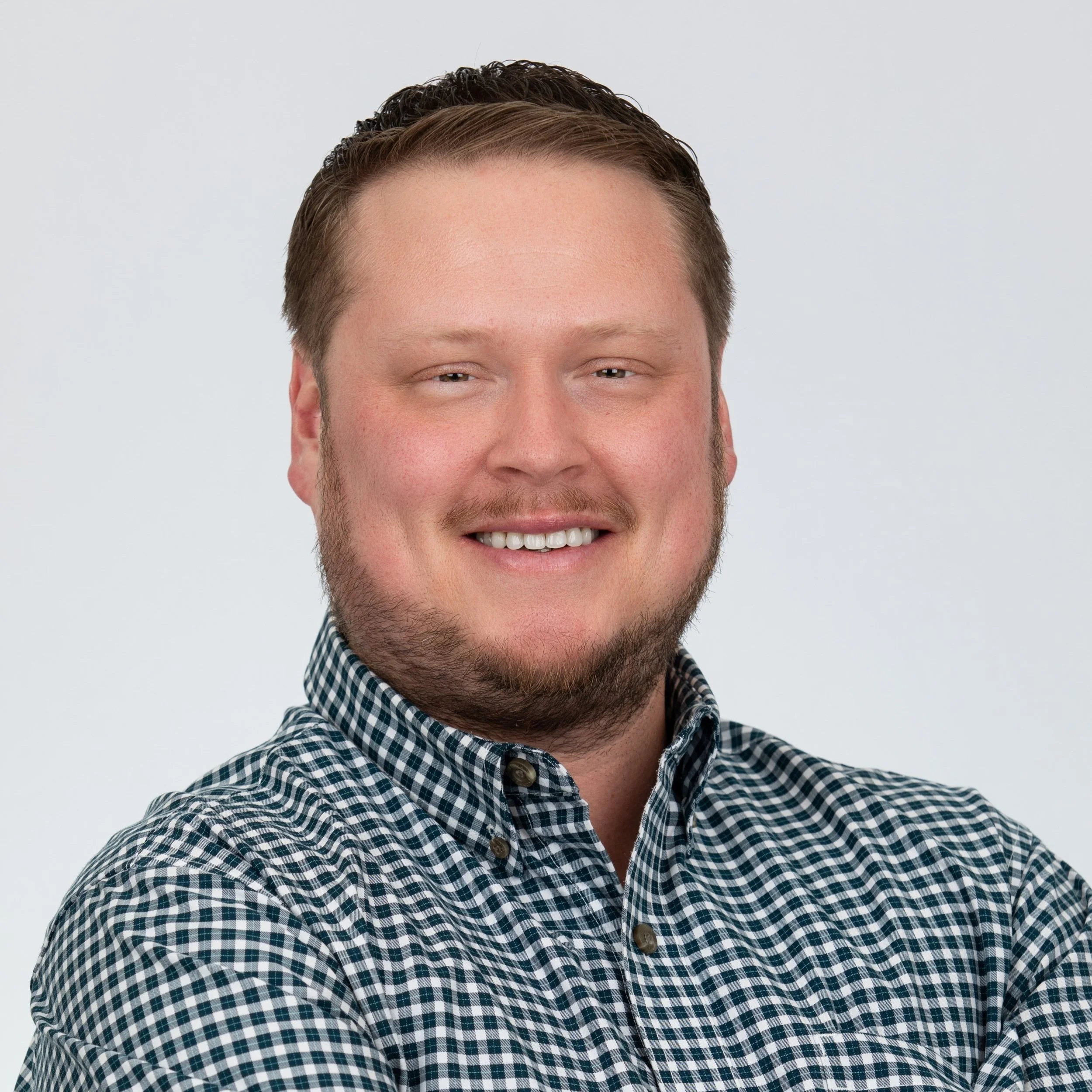 Smiling man with short brown hair and some facial hair wearing a gingham button up collared shirt.