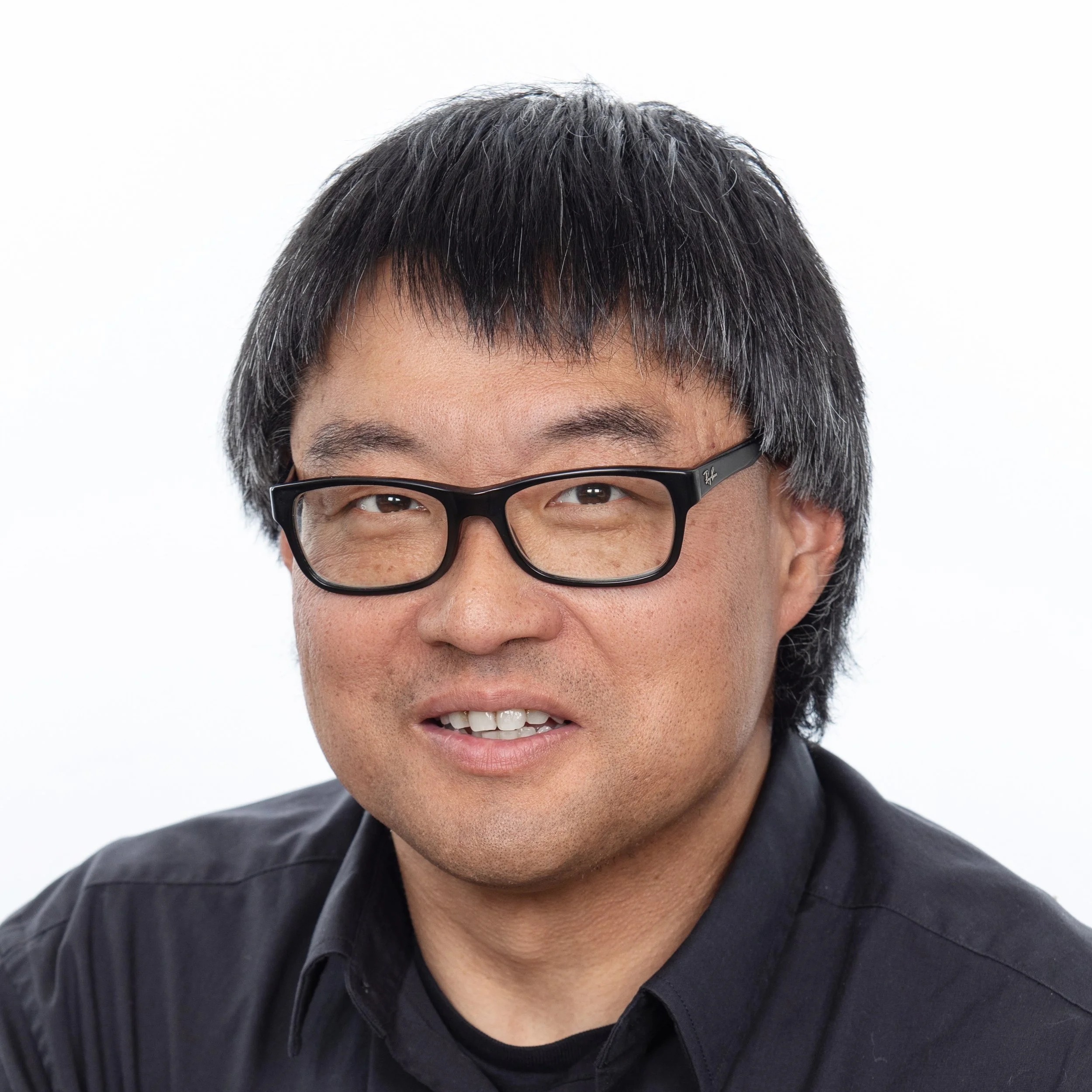A man smiling with dark grey hair and glasses wearing a dark collared shirt.