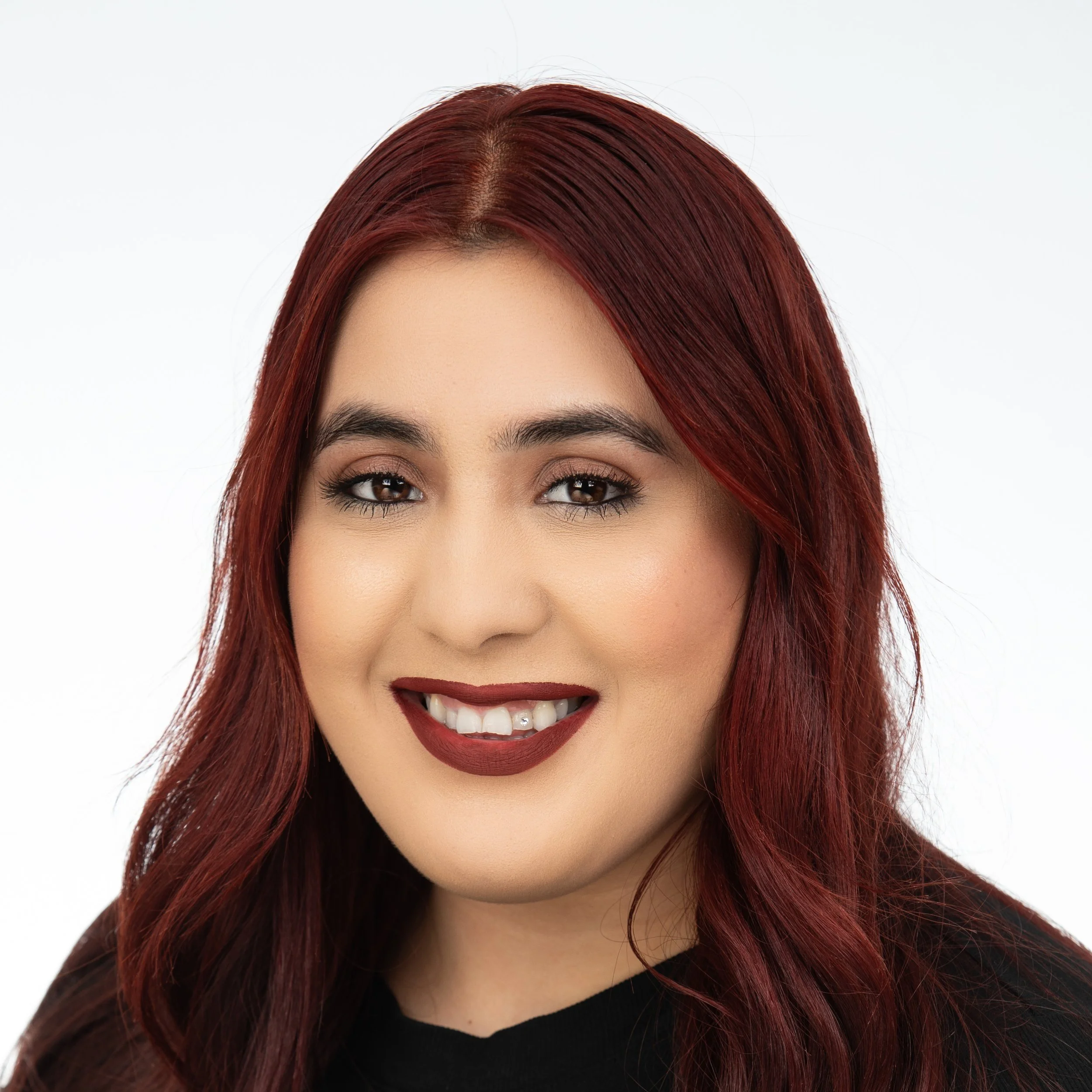 A woman smiling with a tooth gem with dark red long hair wearing a black top.