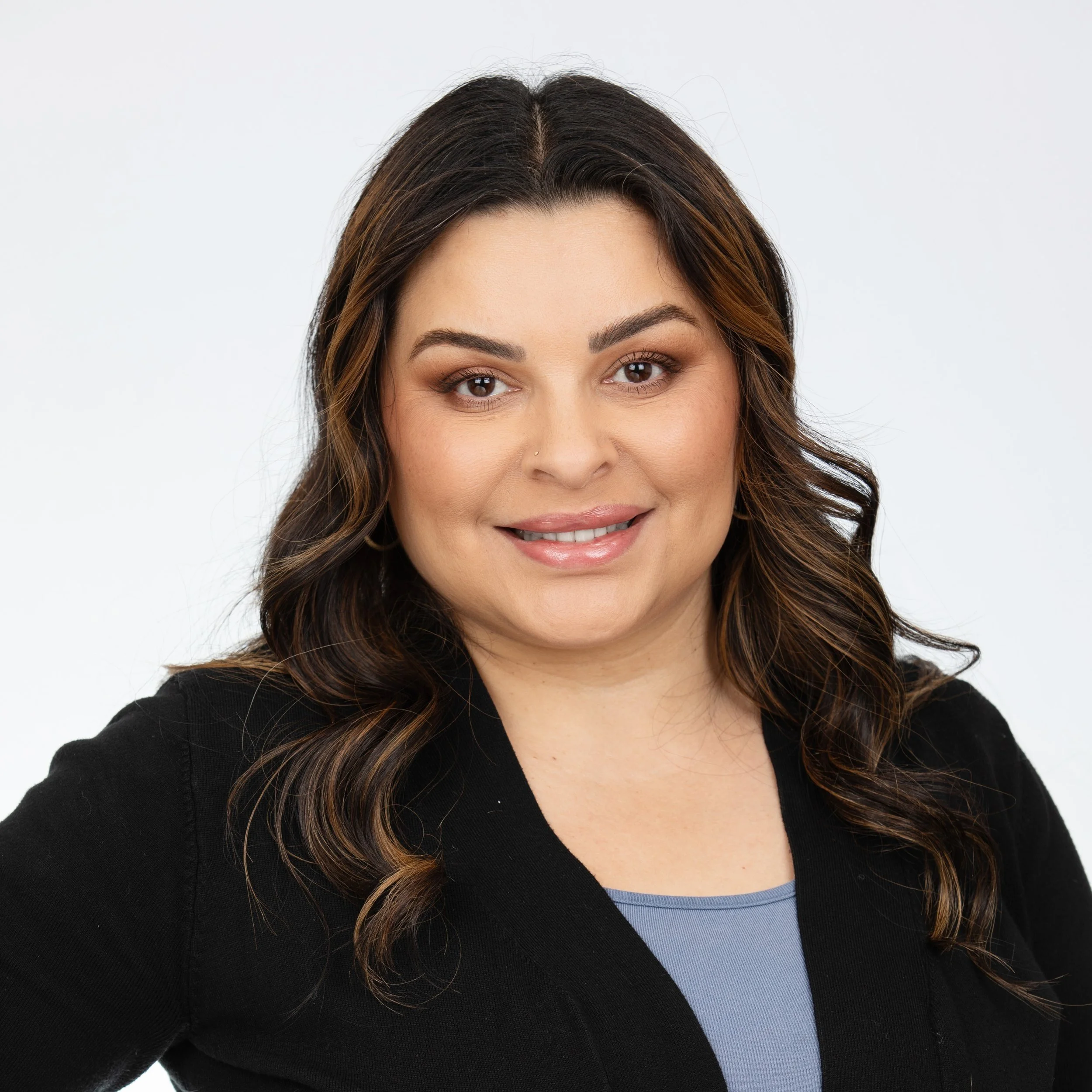A woman smiling with wavy brown hair with low lights wearing a blue top and a black blazer over it.