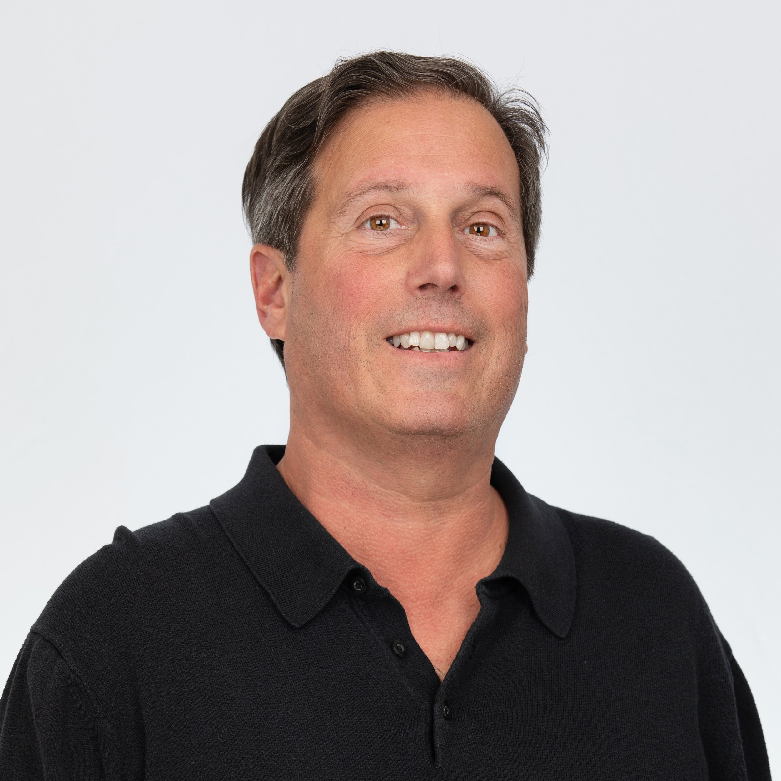 A man smiling with light brown hair wearing a black collared shirt.