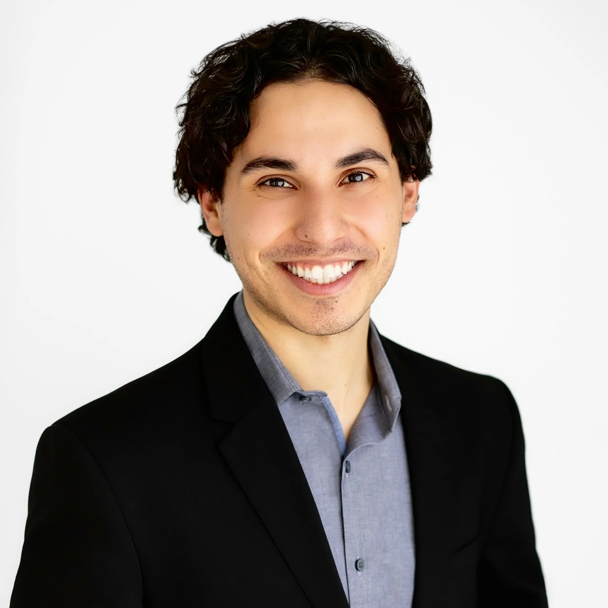 Smiling man with short dark brown hair wearing a blue button up collared shirt and black blazer.