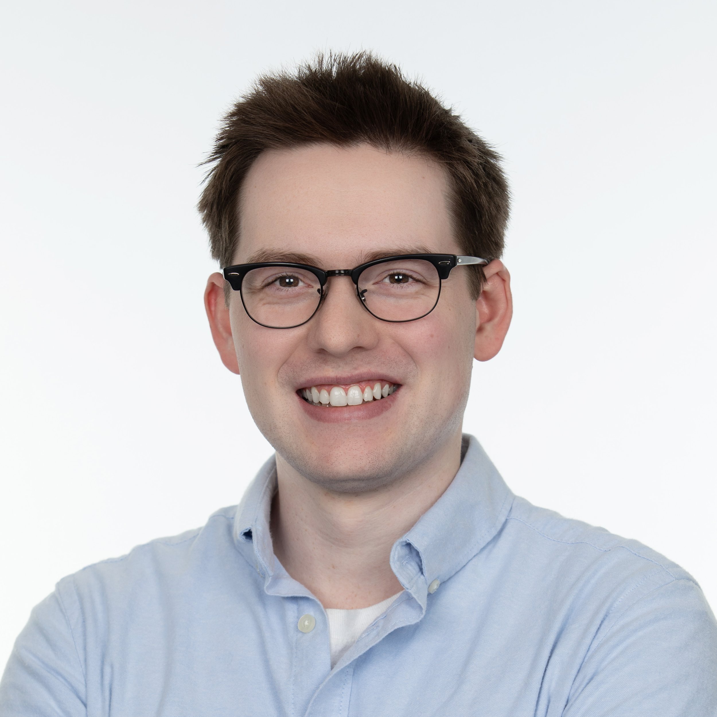 A man smiling with dark brown hair wearing glasses as well as a blue collared button up shirt with a white undershirt.
