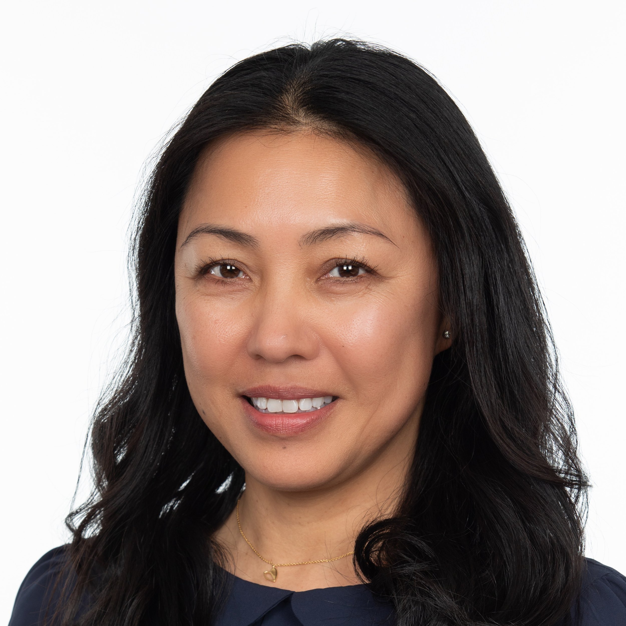 A woman smiling with long dark hair wearing a small necklace with a dark top.