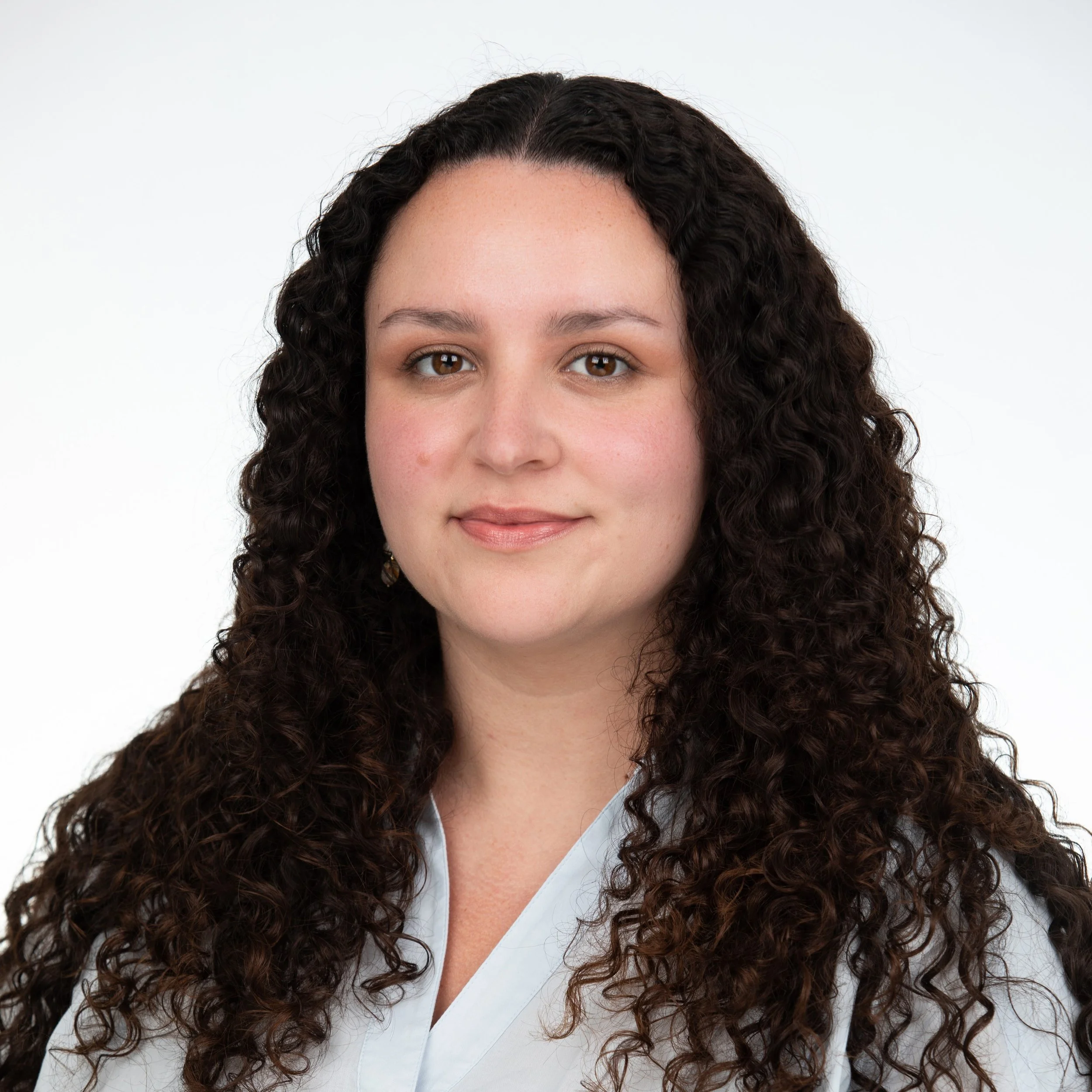 Woman smiling with long dark brown curly hair wearing a light blue blouse.