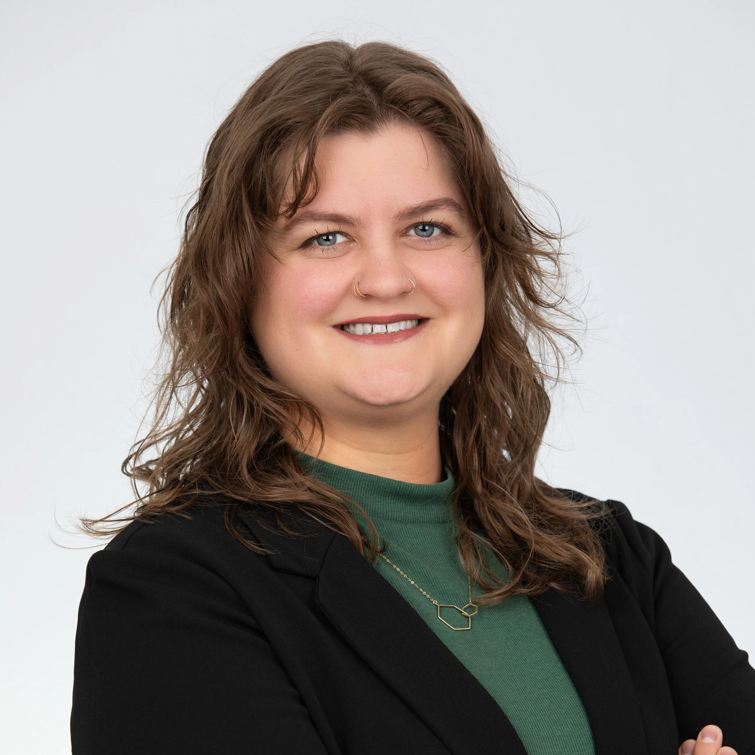 A woman smiling with wavy brown hair wearing a necklace on a green top with a black blazer over it.