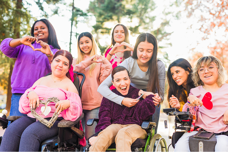 A group of young women and girls outdoors, several in wheelchairs, smiling and making heart shapes with their hands together.