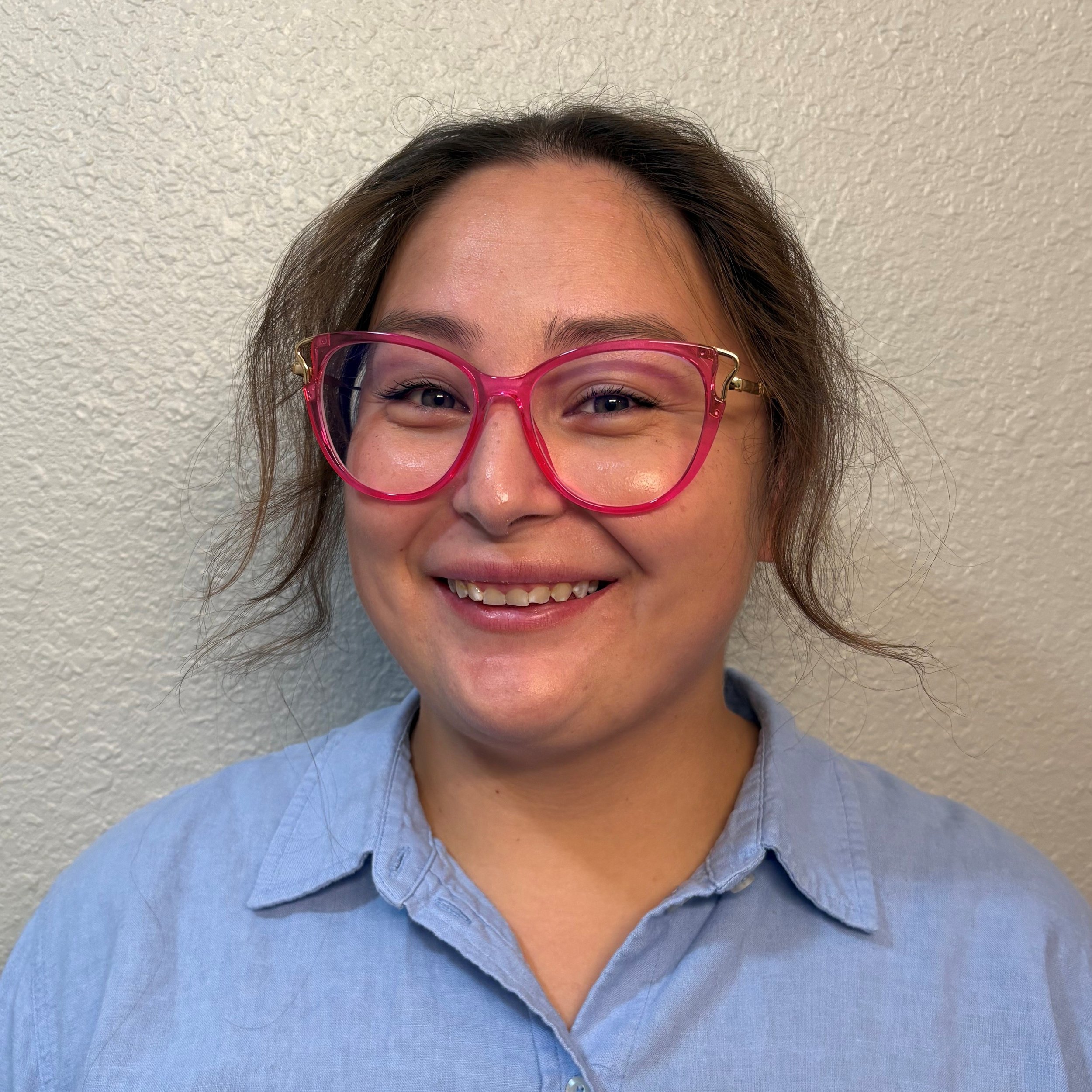 Smiling woman with brown hair pulled back in a ponytail, wearing red glasses and blue collared shirt.