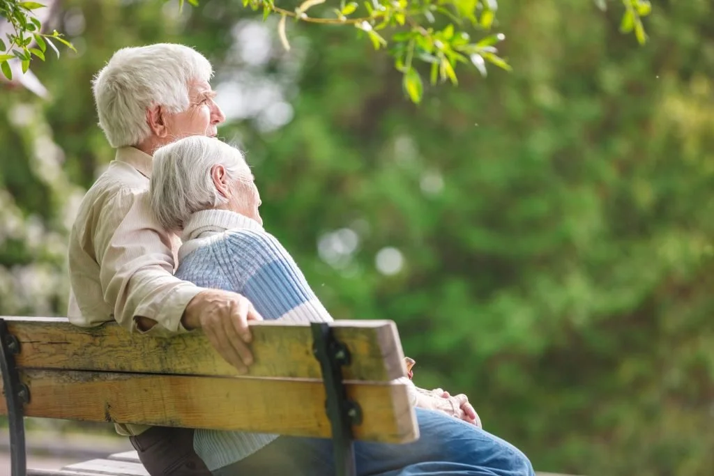 Elderly couple sitting closely together on a bench, enjoying a peaceful moment outdoors with greenery in the background.
