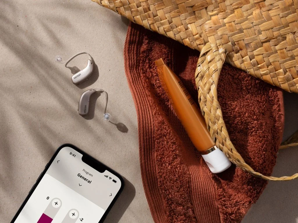 Hearing aids, a phone, orange tube, and a woven bag on a brown towel, all placed on a sandy surface with shadow lines.