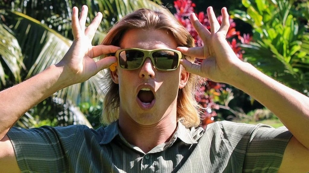 A surprised person with long hair and hay fever eyes wears sunglasses, holding them with both hands among tropical plants outdoors.