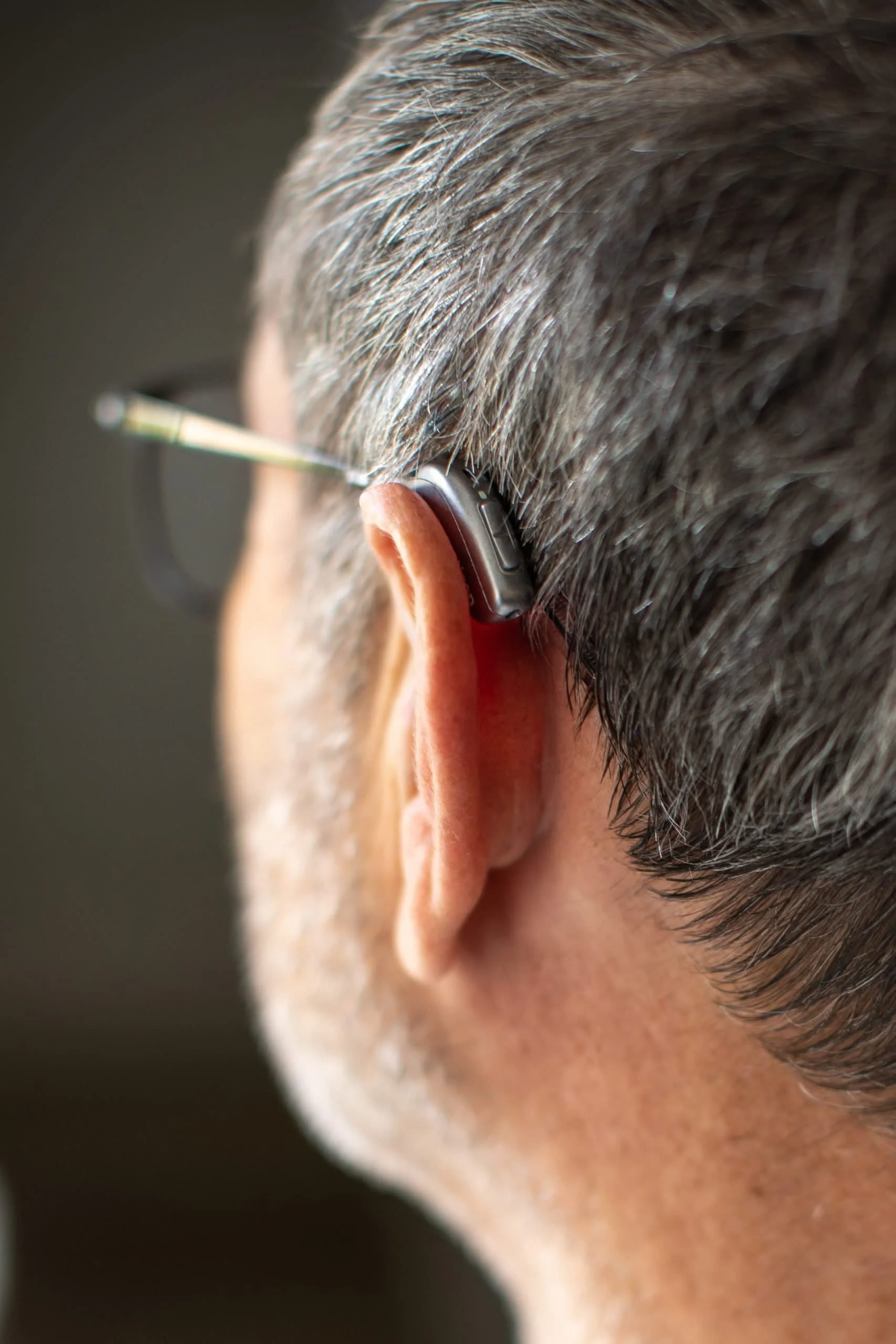Close-up of a mans ear with a hearing aid and eyeglasses, showing short grey hair and part of his face.