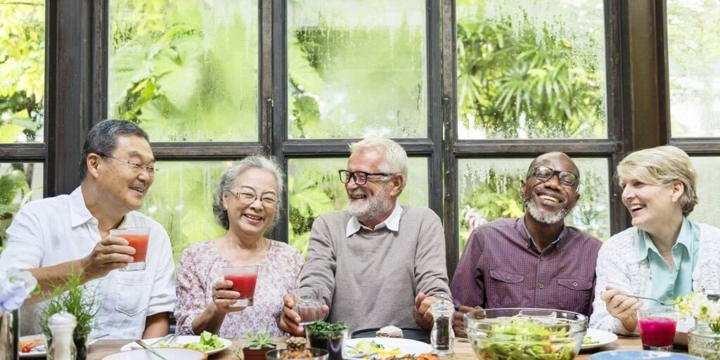 Five seniors sitting at a table, smiling and holding drinks, enjoying a meal together by a large window with greenery outside.