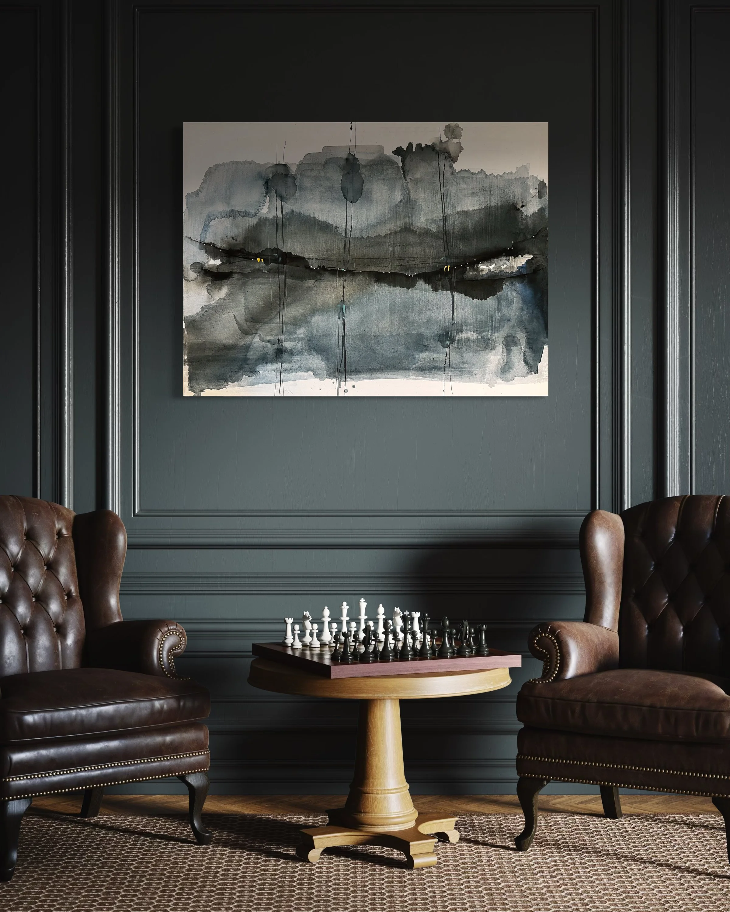 A cozy living room corner with two brown leather armchairs facing each other, a round wooden table with a chess set between them, a geometric patterned rug on the floor, and a large abstract black, gray, and white watercolor painting hung on a dark gray wall with decorative molding.
