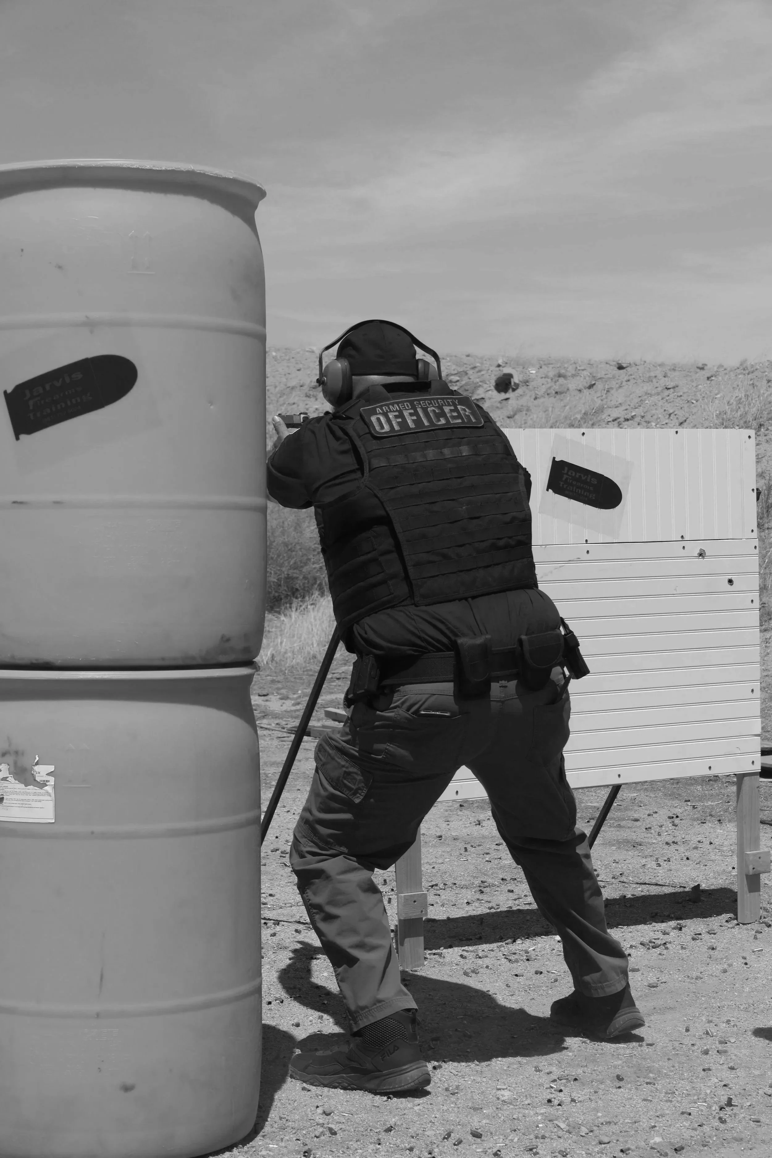 A security officer practicing shooting at a shooting range outdoors, with a large barrel and a shooting target in the background.