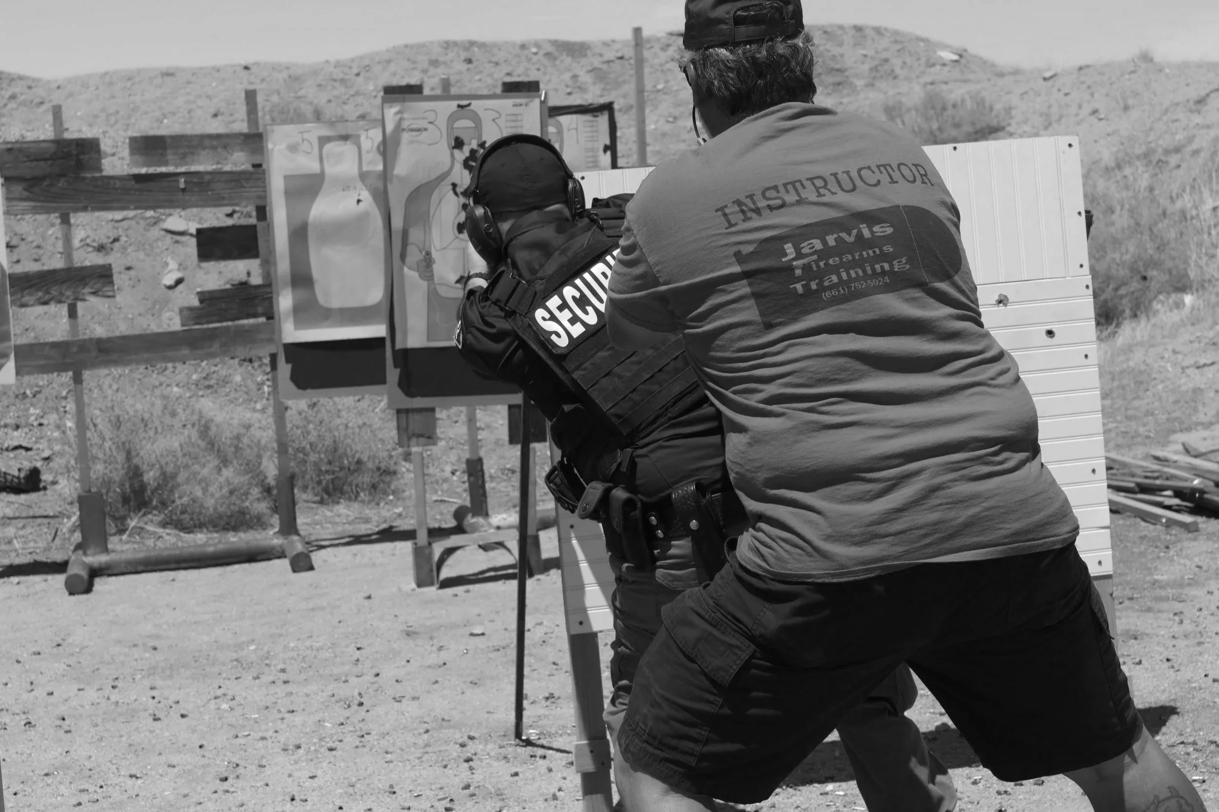 Person in tactical gear with 'Security' patch on arm shooting at targets at outdoor firing range, supervised by instructor wearing 'Jarvis Firearms Training' shirt.