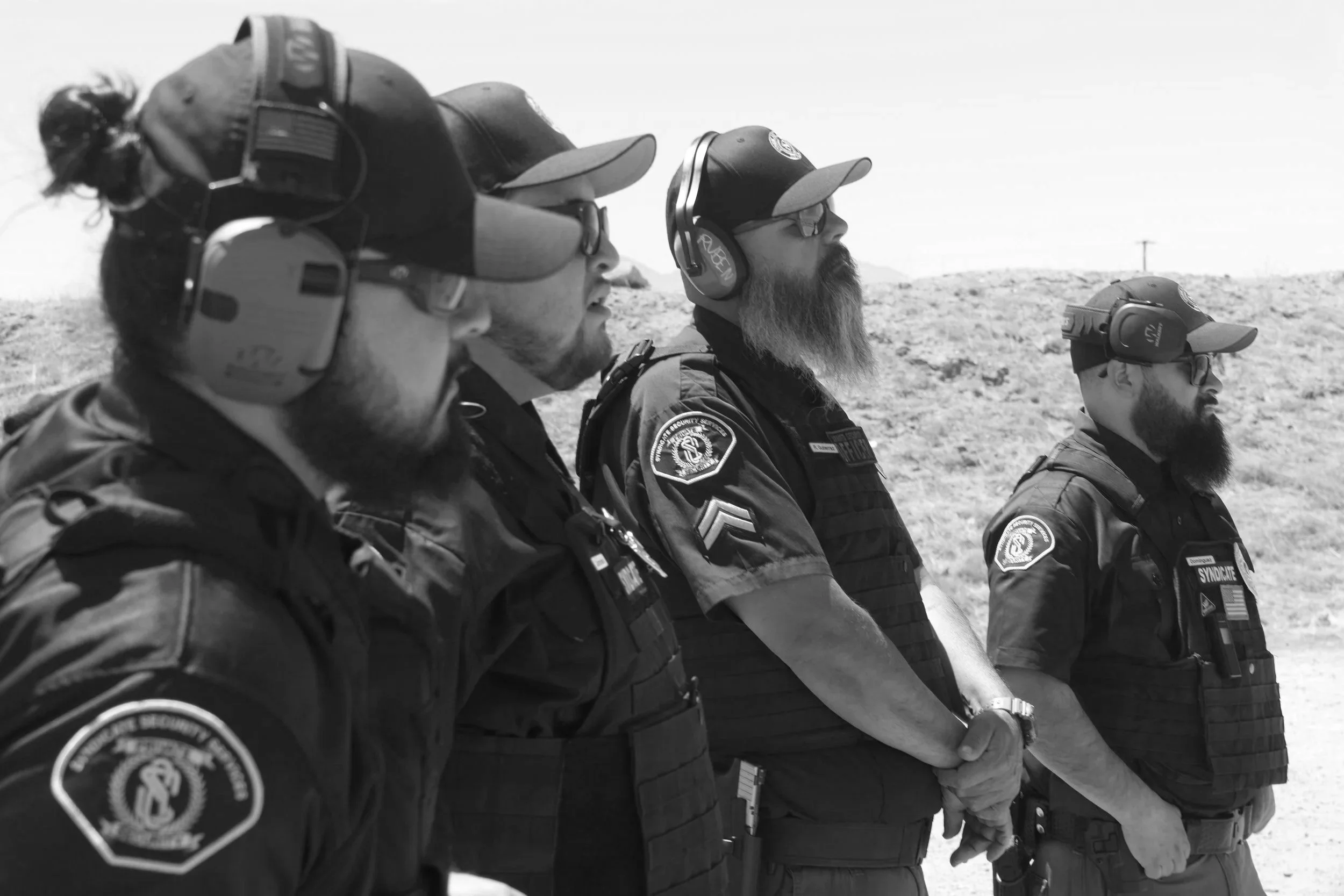 Four uniformed police officers standing in a line outdoors, facing to the right, wearing badges, tactical vests, and protective headgear with earmuffs, in a desert-like landscape.