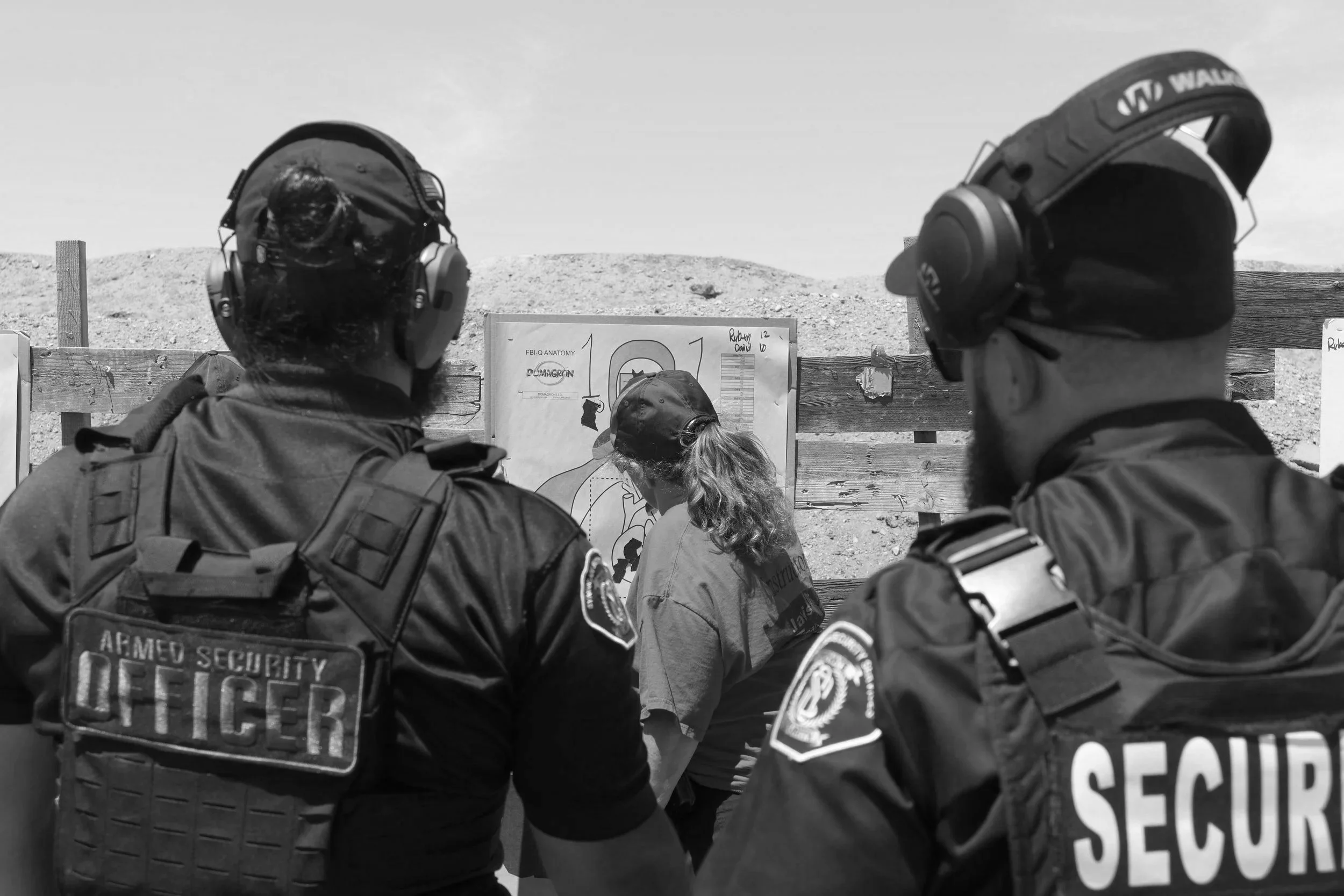 Two armed security officers watching a woman read a background check diagram at an outdoor shooting range.