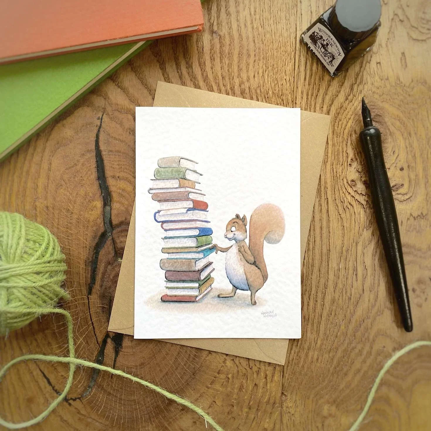 flat-lay-photo-on-a-rustic-wood-surface-showing-an-illustrated-greeting-card-of-a-squirrel-picking-a-book-from-a-big-pile-of-books-by-hannah-nicholls
