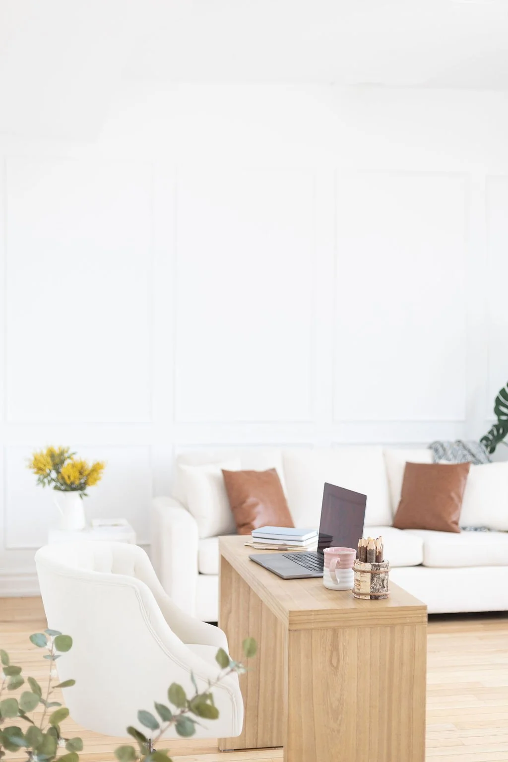 Bright living room with white walls, a white sofa with brown pillows, a wooden coffee table with a laptop, books, a pink mug, and decorated with green plants and yellow flowers.