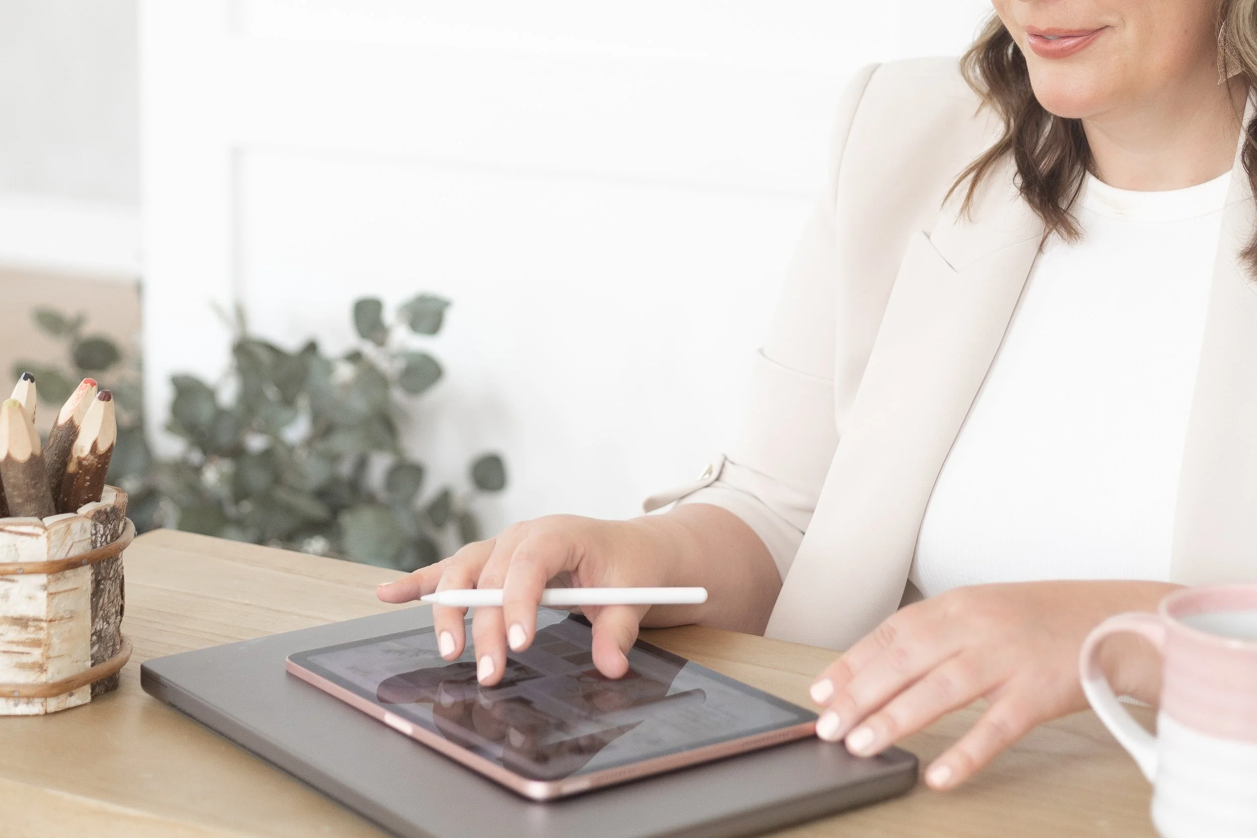 Woman using a stylus on a tablet at a desk with a cup and a container of colored pencils.