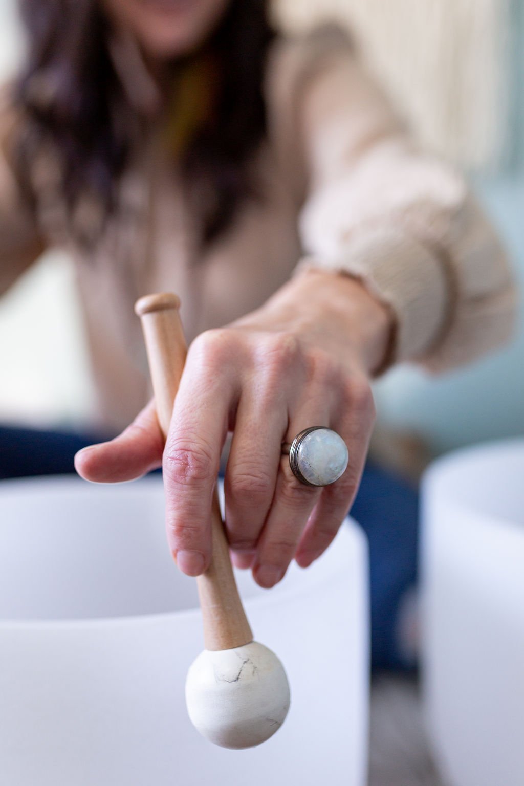 Close-up of a woman’s hand holding a wooden mallet over a white ceramic bowl, wearing a large ring with a moonstone.