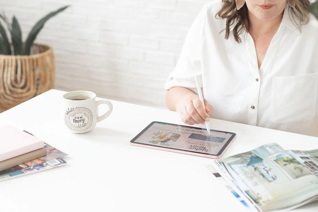 A woman in a white shirt sitting at a white table, writing or drawing on a tablet with a stylus. There is a mug with the words 'Cup of Happy', some magazines, and a plant in the background.