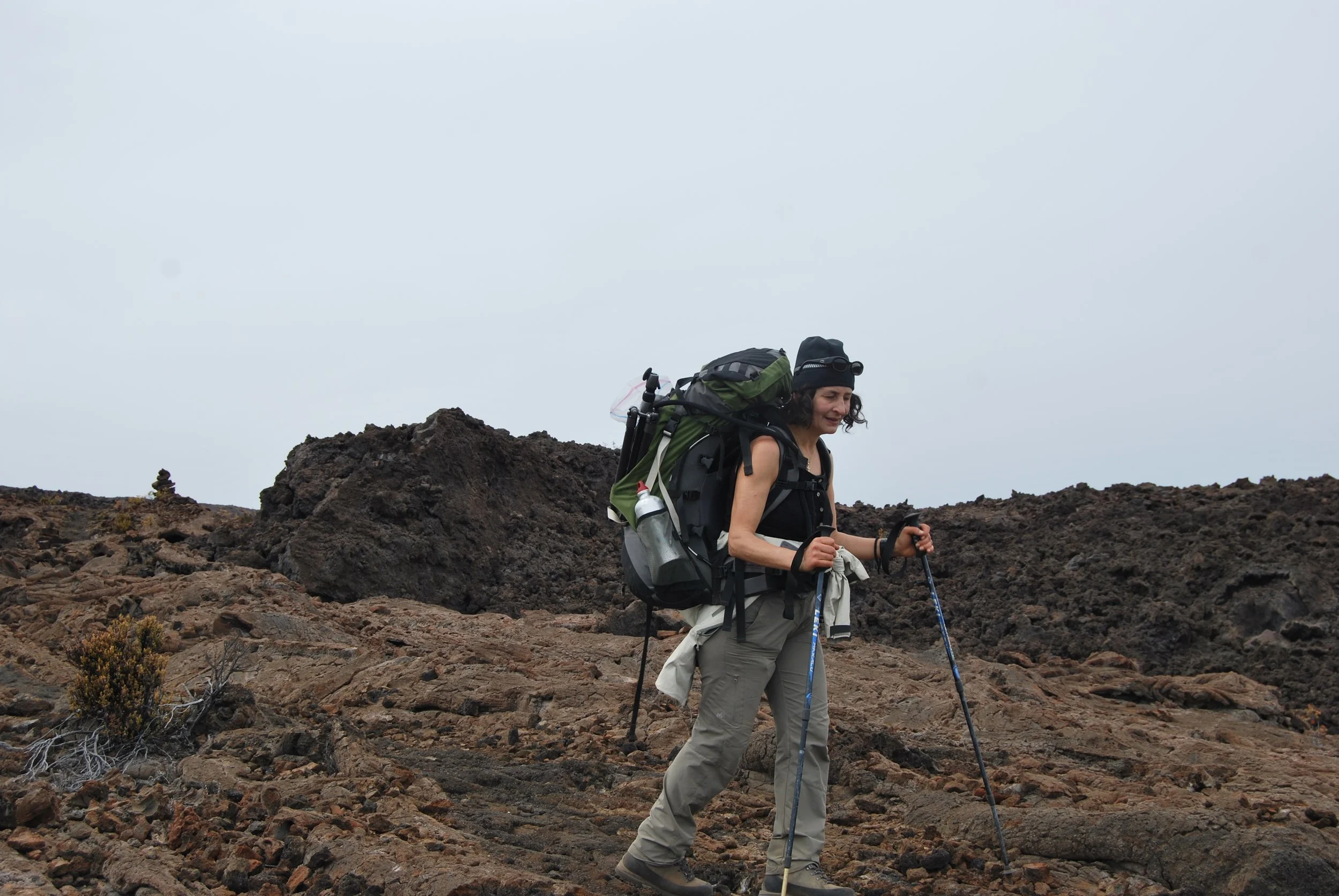 A woman hiking in a rocky, volcanic landscape with a backpack and trekking poles.