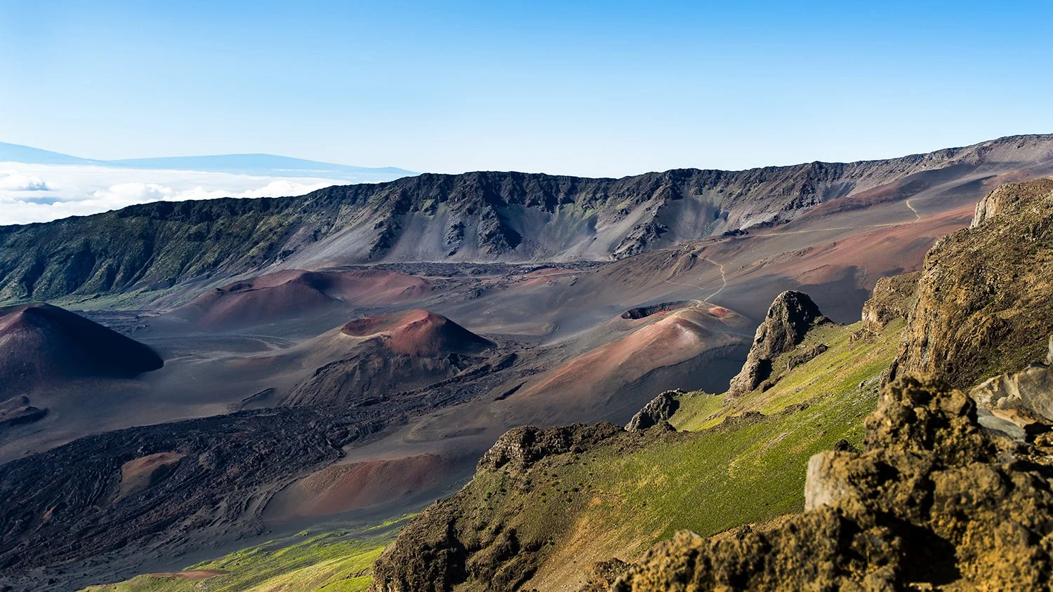 A volcanic landscape with dark volcanic craters, green moss-covered rocks, and distant mountains under a clear blue sky.