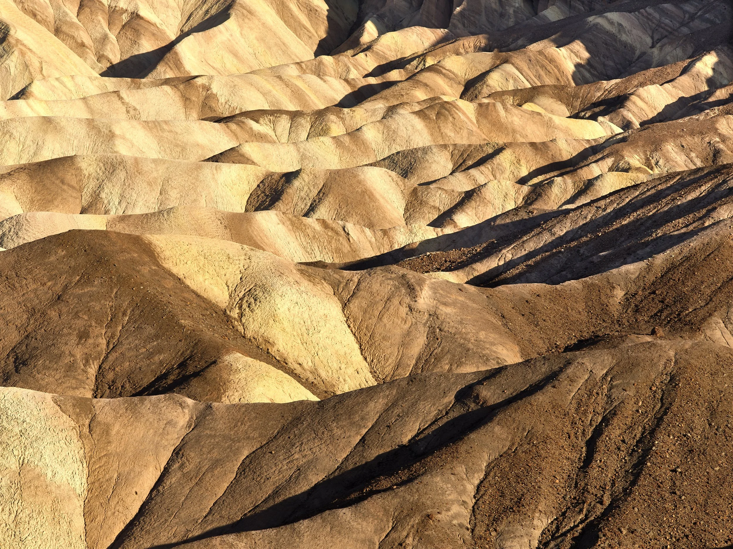 Layered, colorful, eroded rock formations in a desert landscape with shadows cast by the setting or rising sun.