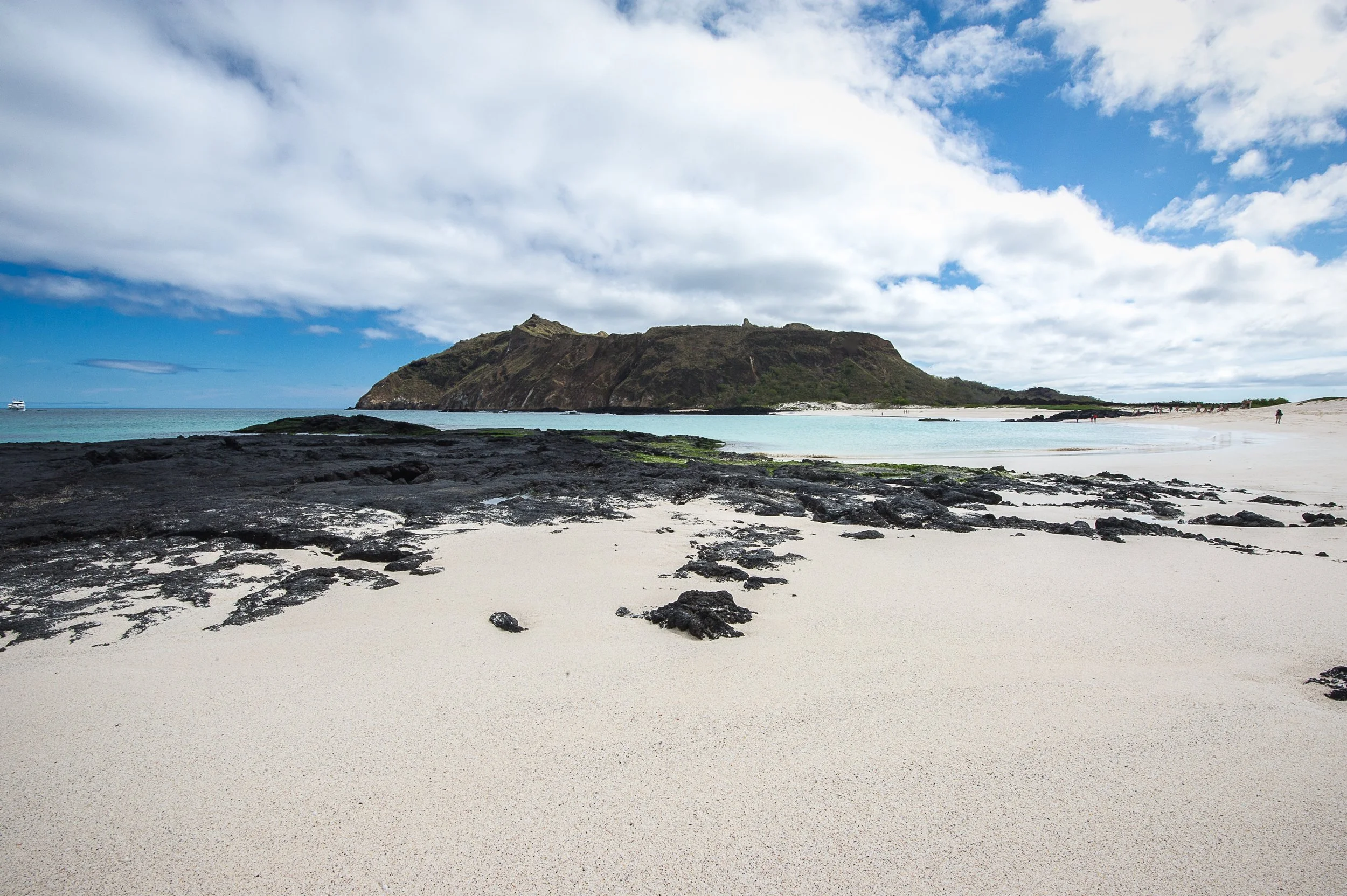 A beach with white sand and rocky areas leading to blue water, with a large grassy hill or mountain in the background under partly cloudy sky.
