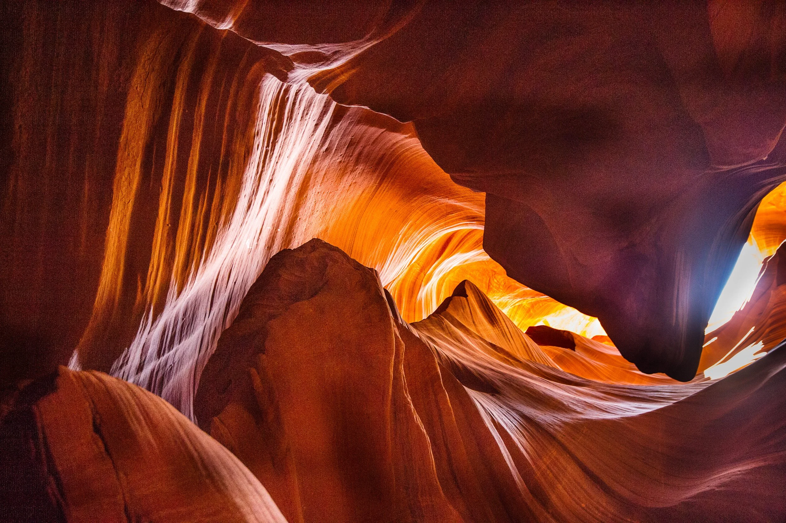 A narrow canyon with smooth, flowing sandstone walls in shades of orange and red, illuminated by sunlight filtering through a small opening.