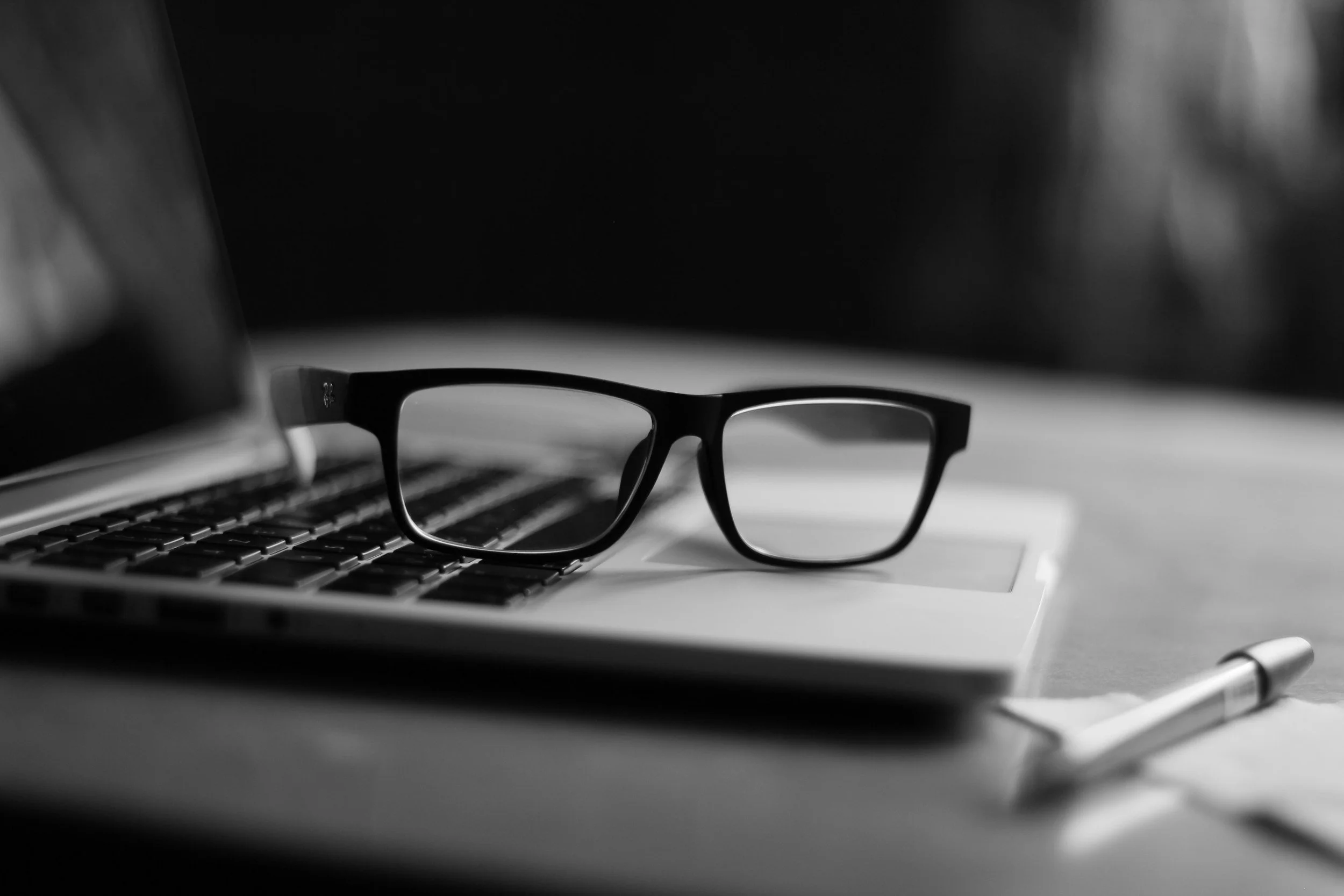 Black and white photo of eyeglasses resting on a keyboard, with a pen and a laptop or notebook nearby.