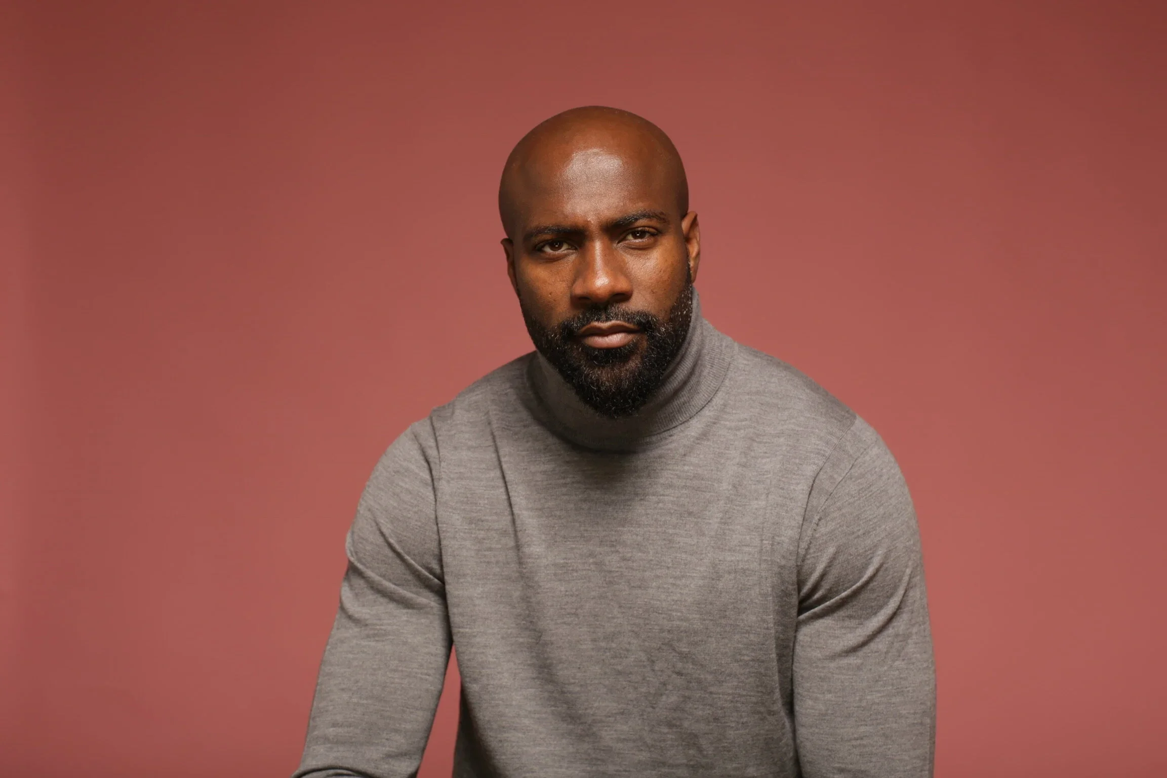Portrait of a bald man with a beard wearing a gray turtleneck sweater against a pink background.