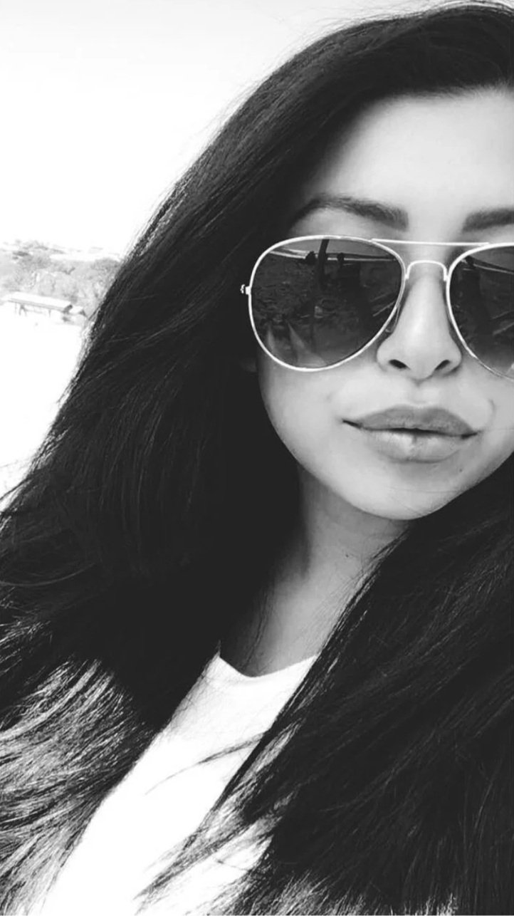 A black and white photo of a woman with long dark hair wearing sunglasses, with a slight smile, outside in a sunny environment.