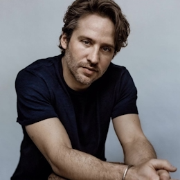 A man with brown hair and a beard wearing a black t-shirt, posing with his arms crossed against a light gray background.