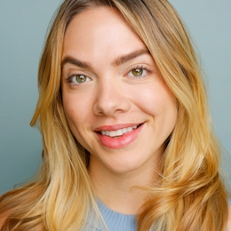 Close-up of a young woman with blonde hair and green eyes smiling against a blue background.