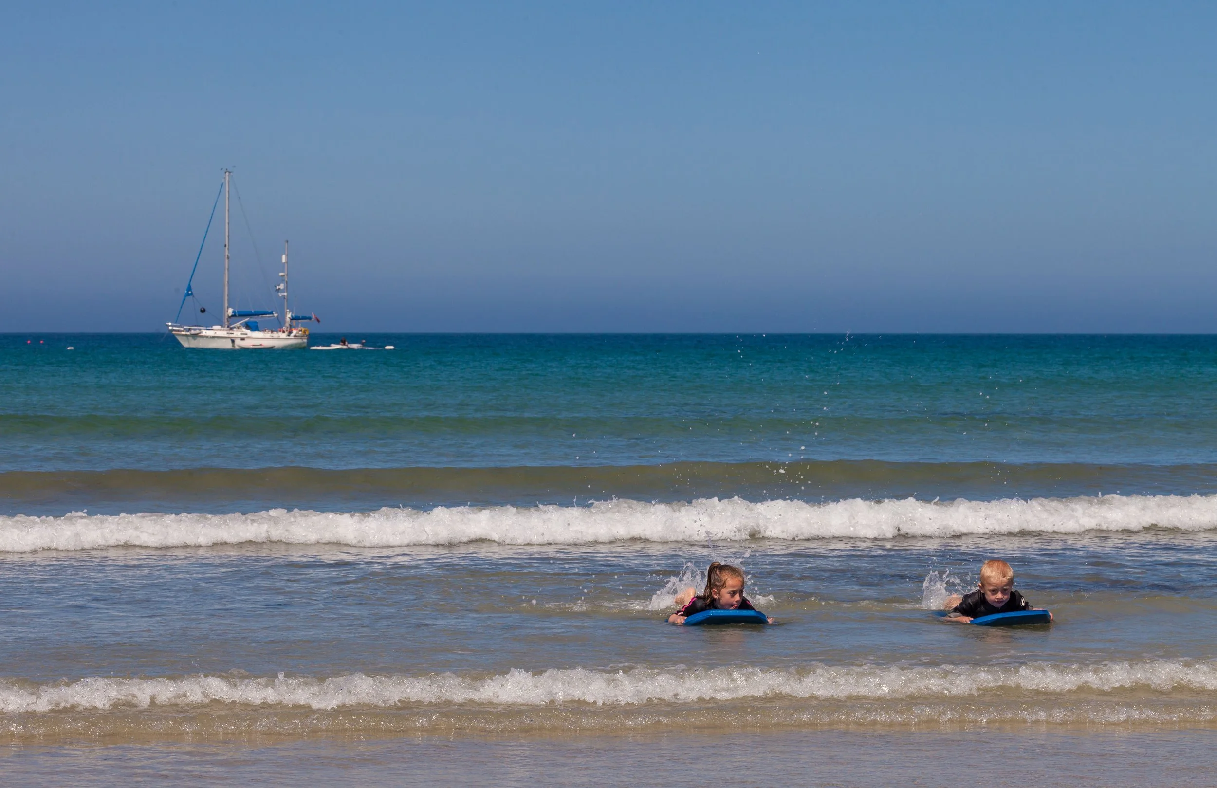 Pembroke bay, with a sailboat on the horizon and children body boarding in the sea.