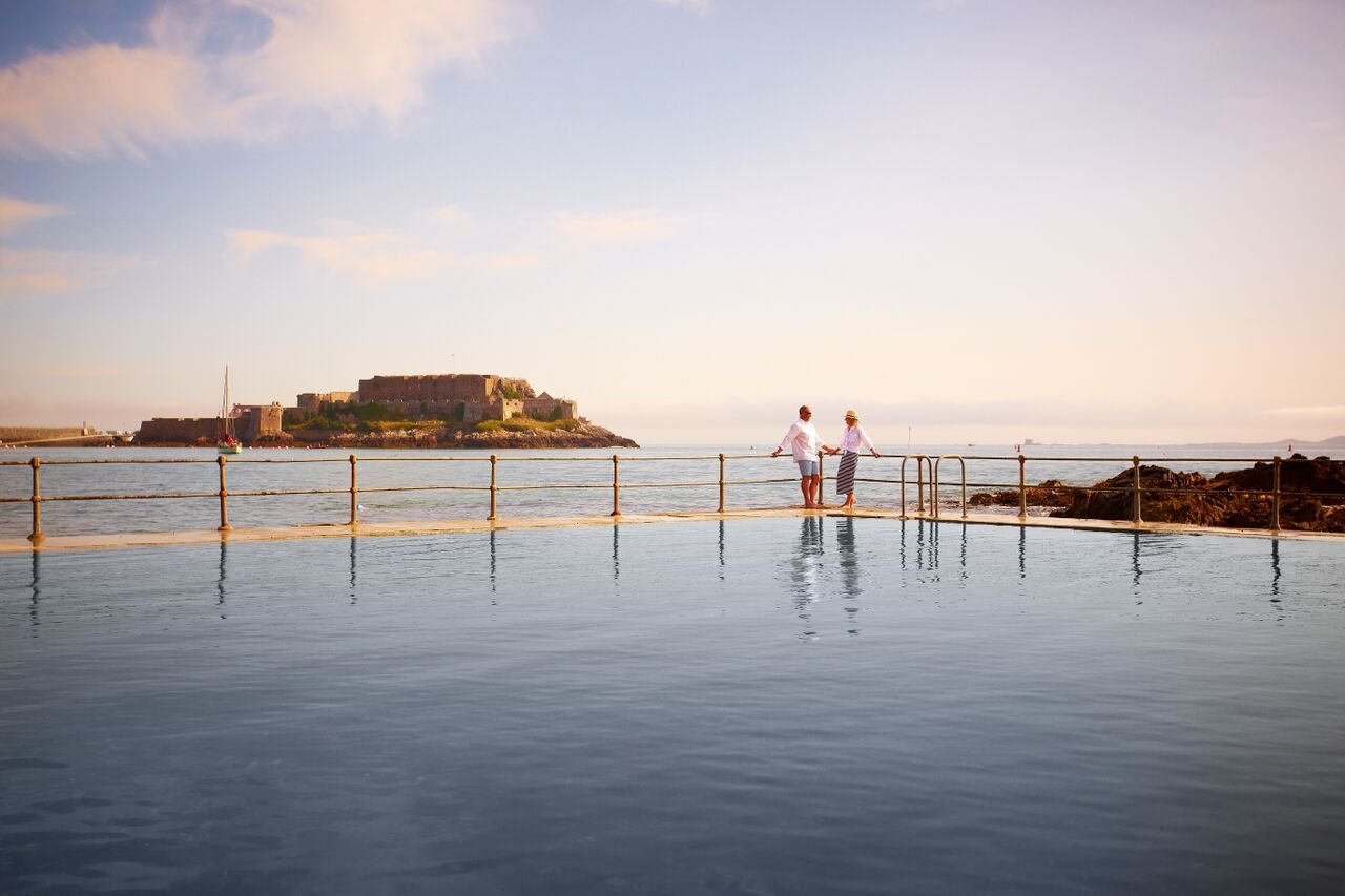 Couple standing by railings at La Vallette Bathing Pools, high tide with Castle Cornet in background, Guernsey