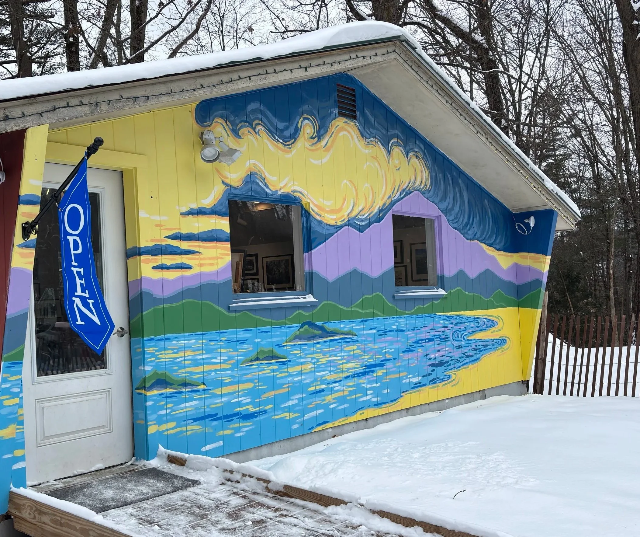 Colorful exterior mural of a lake scene with mountains, clouds, and sky painted on the side of a building, with a snow-covered ground in front and a blue 'Open' flag hanging near a door.