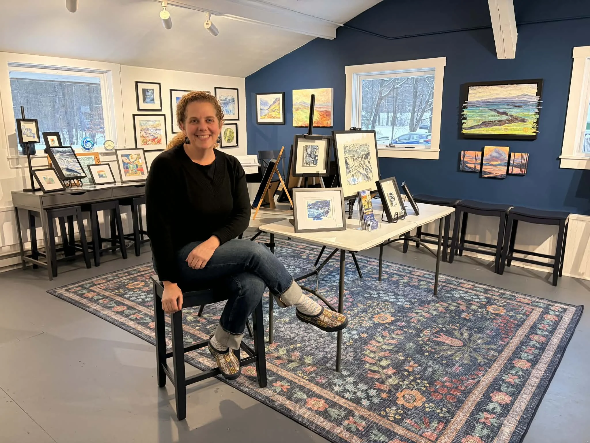 A woman with curly hair sitting on a black stool inside an art gallery with framed paintings on walls and tables, a colorful rug on the floor, and windows showing snow outside.
