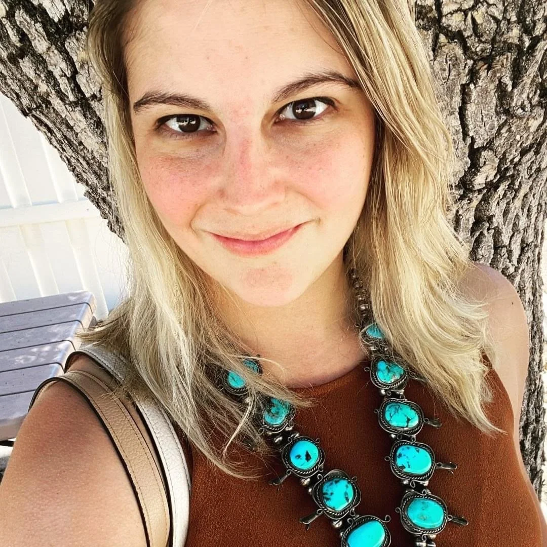Close-up of a young woman with blonde hair taking a selfie outdoors, smiling, standing in front of a large tree. She is wearing a brown top and a turquoise jewelry necklace.
