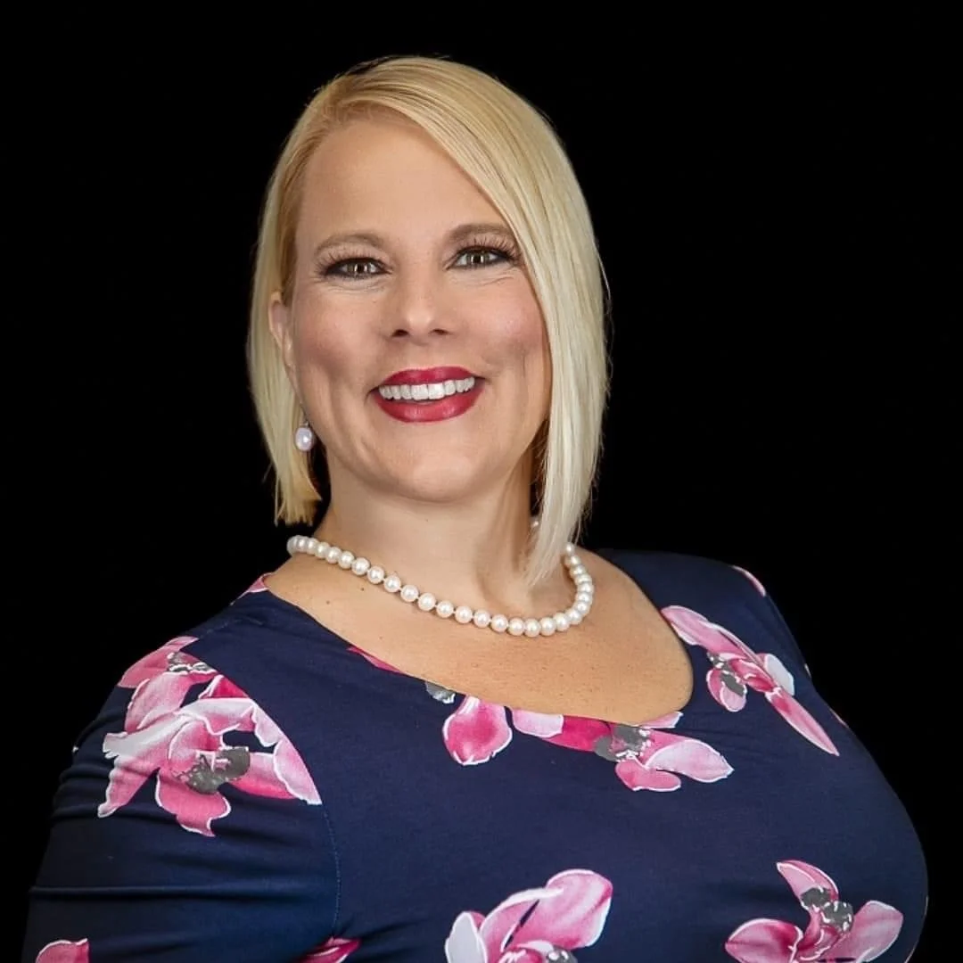 A woman with blonde hair, red lipstick, pearl necklace and earrings, wearing a navy blue dress with pink floral pattern, smiling against a black background.