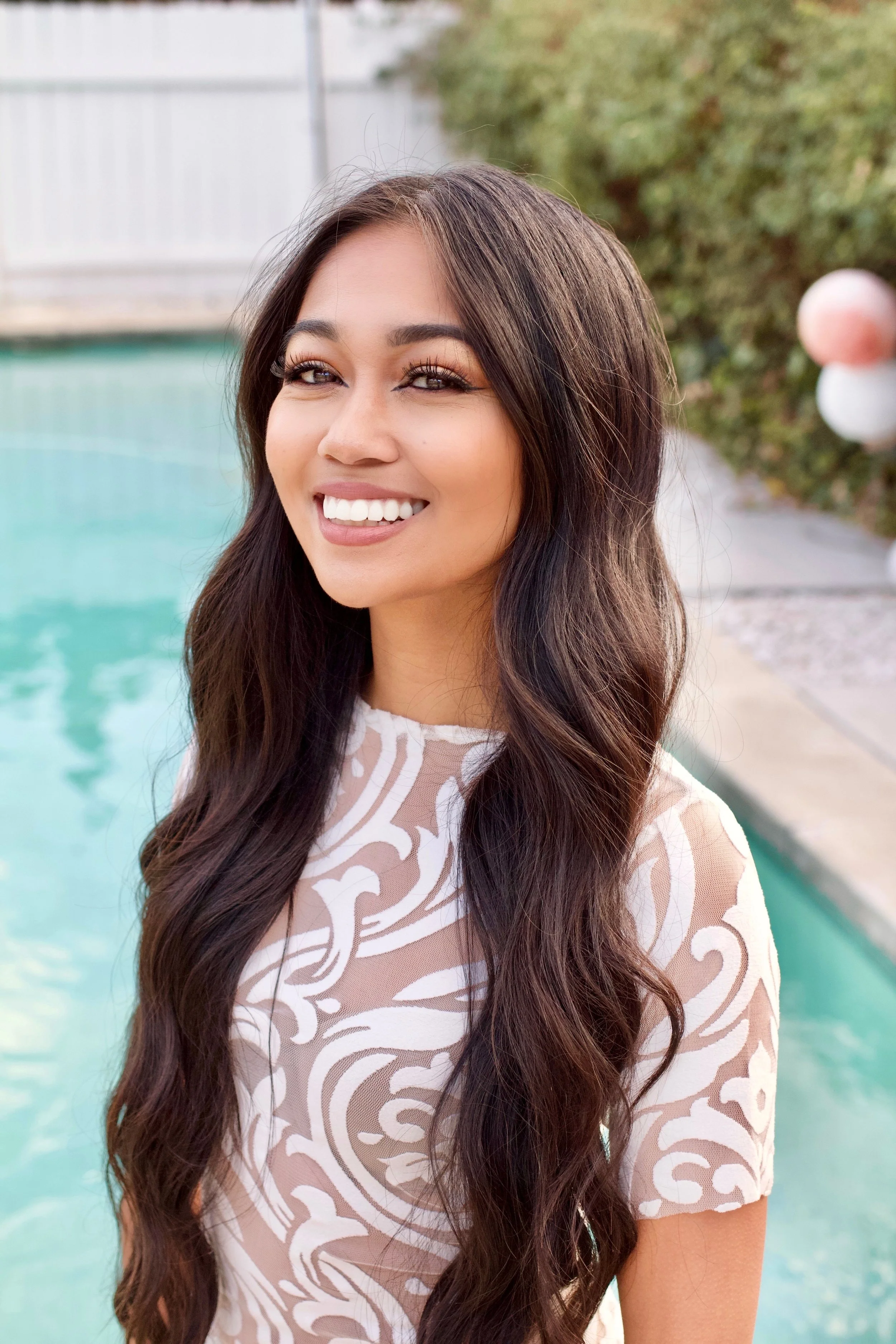 Portrait of a smiling young woman with long wavy dark hair, wearing a beige and white patterned top, standing outdoors next to a swimming pool.