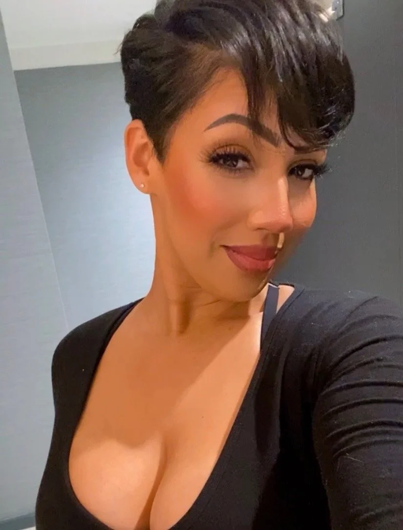 A woman with short dark hair and makeup, wearing a black top with a deep neckline, taking a selfie indoors.