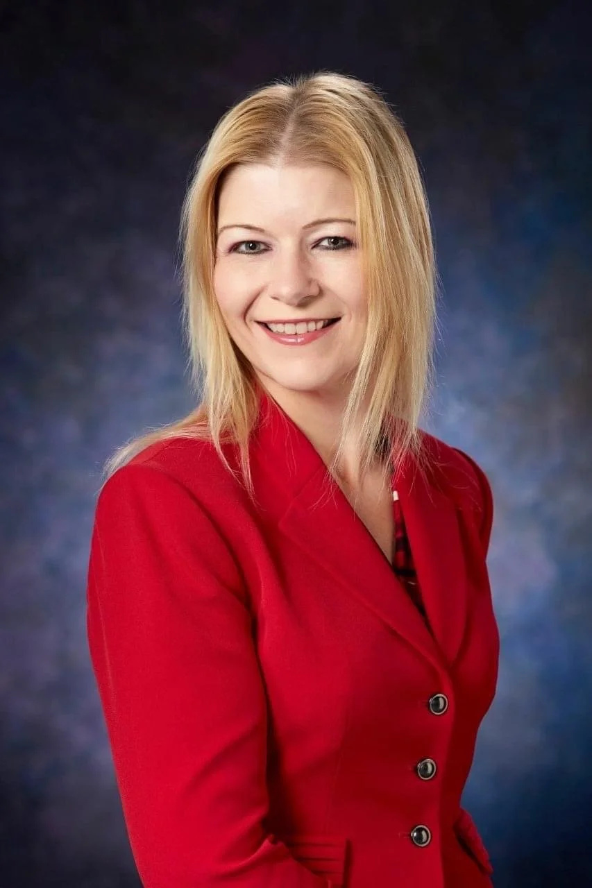 Professional woman with blonde hair smiling, wearing a red blazer, against a dark blue background.