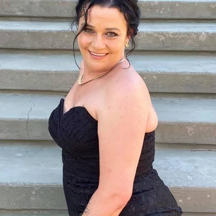 A woman in a black strapless dress, smiling and sitting on concrete steps outdoors.