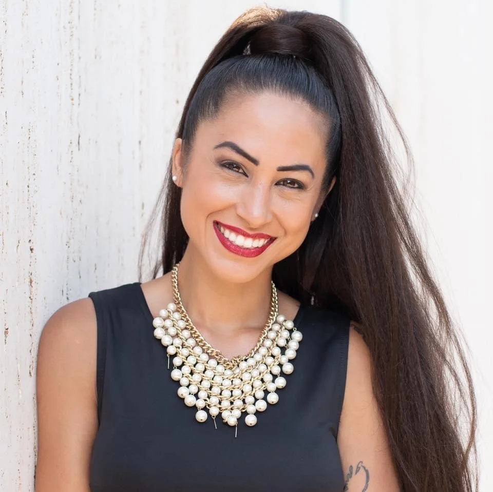 A woman with long dark hair styled in a high ponytail, wearing a black sleeveless top, a layered pearl necklace, earrings, and red lipstick, smiling in front of a white wooden background.