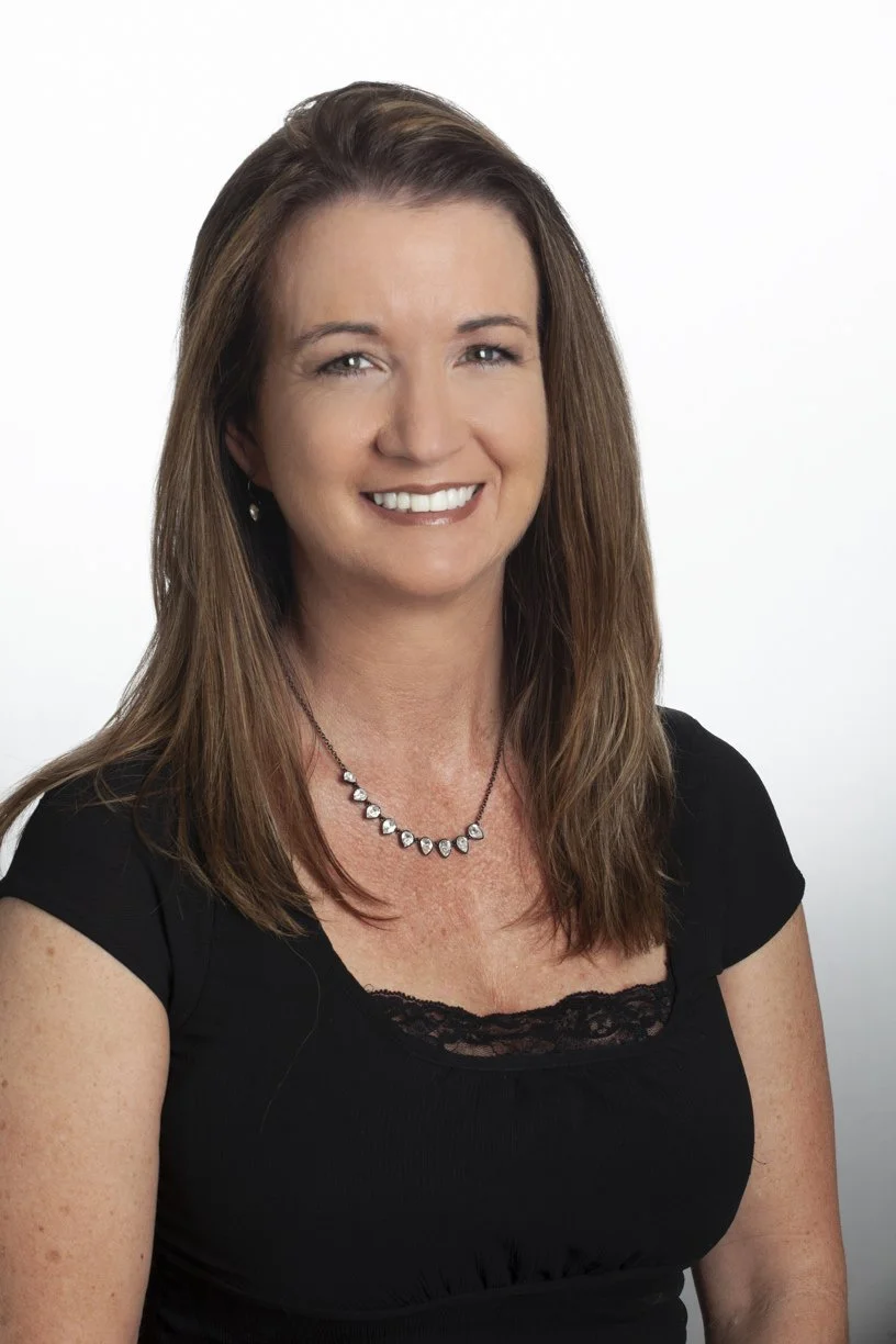 A woman with shoulder-length brown hair, wearing a black top with lace detail, a diamond necklace, and earrings, smiling at the camera against a white background.